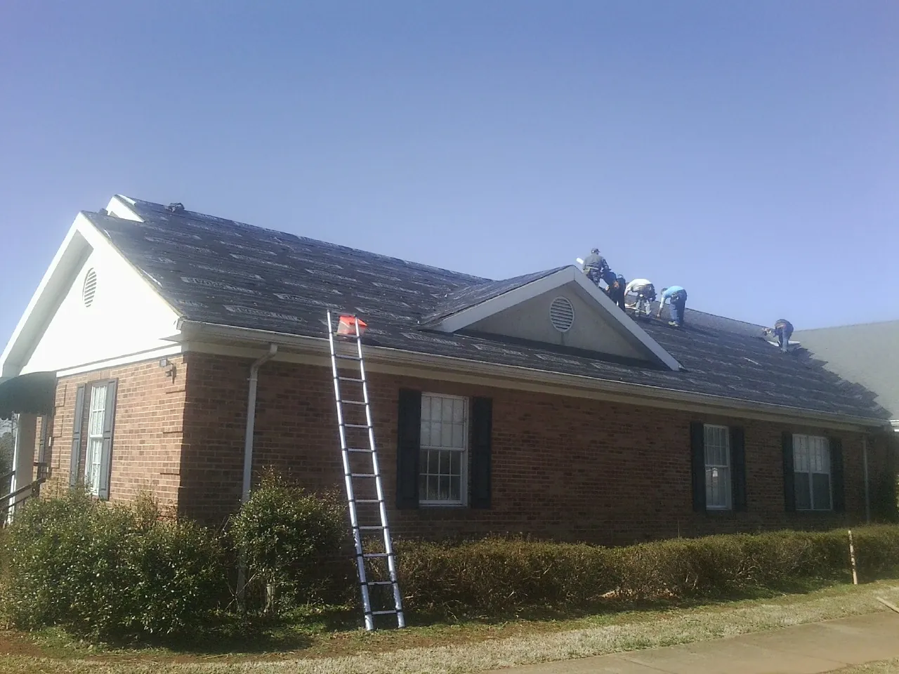 Crew tearing shingles off a brick ranch roof with a ladder set — Metal Roof Repair work by our crew in Glasco